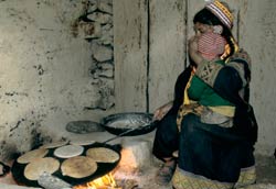 Woman squats, toddler on her lap, cooks large pan full of flat bread over indoor cookstove, flames visible beneath pan