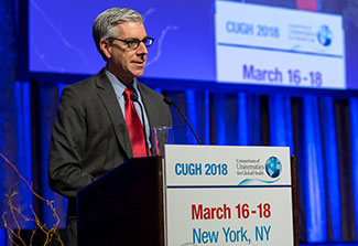 Dr. John Balbus speaks from a podium labeled CUGH 2018, slide projected in the background