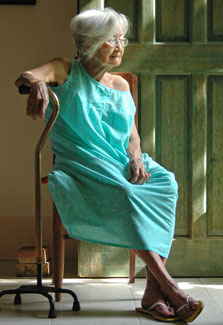 Frail looking elderly woman wearing a house dress seated in doorway leans on a cane
