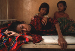 emaciated woman lying down in old hospital bed, two young children look on from behind