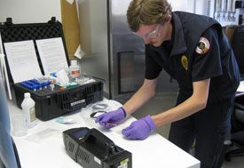 Man leans over table, wears purple disposable gloves and protective glasses, performing test, large case with many bottles