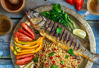 Image of grilled fish plated along side cuts of tomatoes, bell peppers, couscous, greens and lime