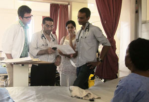 Man holds medical chart, three people look on, two wear stethoscopes, patient sits in foreground next to empty hospital bed