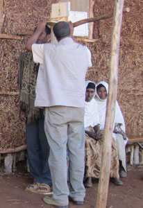 Man measures the height of another man, standing, women seated in background, outdoors next to a rough hut