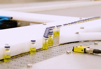 Small glass vials filled with yellow liquid moving along an industrial conveyor or filling line, guided by white rails