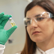 Female researcher with safety glasses on, holding up and observing a vial in her green gloved hand