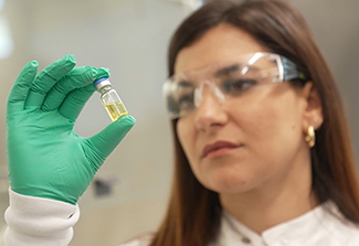 Female researcher with safety glasses on, holding up and observing a vial in her green gloved hand