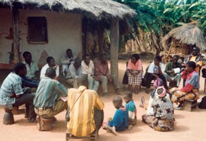 Large group of adults and children, seated on stools and dry dusty ground in circle, huts in background