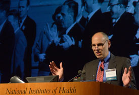 NICHD Director Dr. Alan E. Guttmacher speaking at a podium, slide projected in background