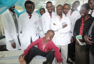 Smiling, health-looking young African boy in hospital bed surrounded by many healthcare workers