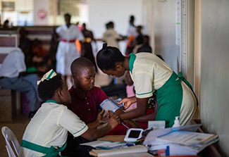 The photo on this page is set in a health care facility and shows two female professionals,wearing pale yellow and green uniforms, adjusting the blood pressure cuff for a male patient wearing ared shirt.