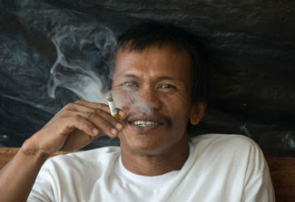 Indonesian man holding smoking cigarette smiles at the camera, puffs of smoke visible