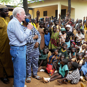  In this photo, President Jimmy Carter, wearing a blue shirt, speaks into a microphone and addresses a crowd of Ghanian children outside a hospital 