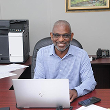 John Lindo smiling while sitting at a desk, typing behind a laptop