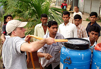 In Dhaka, Dr. Eric Nelson pumps water through a hose from a pond into a blue barrel, surrounded by others who are helping