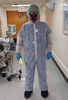 Dr. Mark Brady stands in the hallway of a hospital wearing full PPE, including a hair net, half-mask respirator, suit gloves.