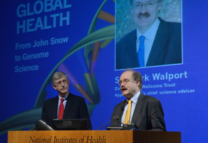Sir Mark Walport speaks at a podium, NIH Director Francis S Collins looks on, slide reading global health projected