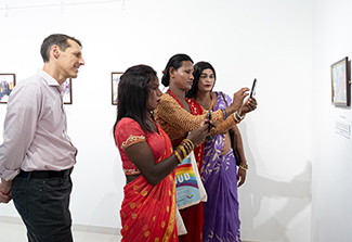 In this photo, U.S. Embassy, Nepal, Chargé d'affaires Jason Meeks, wearing a striped pink shirt and black pants, watches as three trans women in colorful Nepalese dresses take a selfie at the Sweekar Photo Exhibit at Siddhartha Art Gallery in Nepal. The exhibition features photos taken by rural trans women from the Madhesh Province, reflecting their experiences in connection with their gender identities and in obtaining HIV care.