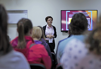 This photo is taken at the back of a classroom so that you see the backs of students’ heads as they focus on Dr. Melinda Gal, wearing a dark sweater and pink blouse, while she lectures in a classroom in Cluj, Romania.