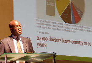 Dr. Nelson Sewankambo speaks into microphone at podium, slide reads 2,000 doctors leave country in 10 years
