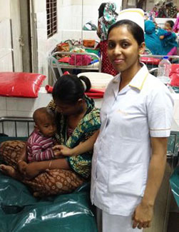 Nurse stands next to a bed, where a mother comforts a baby, in a crowded hospital ward