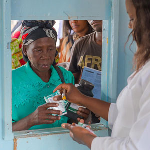 Medical worker in white coat distributes medications through a service window to an older woman