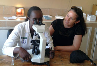 In lab Dr. Wendy Prudhomme O’Meara helps Kenyan researcher, who is looking in a microscope