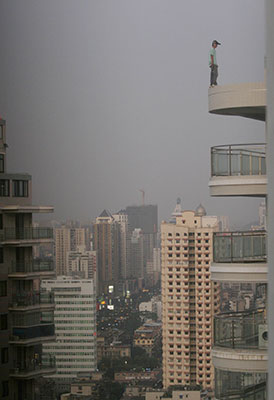 Person stands precariously on edge of balcony on very high floor of tall building, city skyline with tall buildings in background