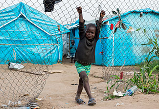 Young boy hangs on a chain link fence in front of tents in a refugee camp.