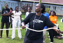 African woman in foreground in athletic wear holds rope, whistle around her neck, more women in background also wearing athletic