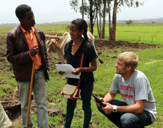 Two researchers collect data on tablets while speaking with a man in front of a field on a farm, cow in background