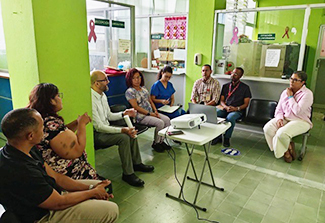 A group of eight people seated in a semicircle inside a green-walled clinic or health center, gathered around a small table with a laptop and projector, appearing to be in a meeting.