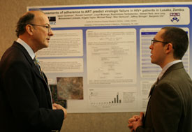 Dr Roger I Glass speaks with man in front of poster pinned to large bulletin board, displayed in conference room