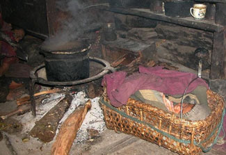 Small baby wrapped in blankets in small basket on floor of a dark hut, very close to smoking wood burning fire