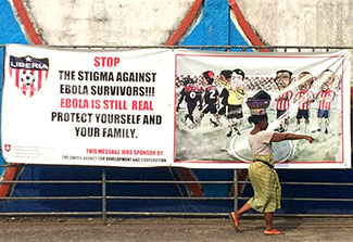 Woman walking in street in Monrovia, Liberia, in front of large football banner reading: stop the stigma against Ebola survivors