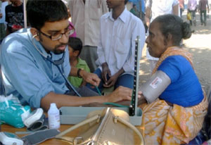 A male medical worker with stethoscope takes the blood pressure of an older woman, seated, in an outdoor clinic