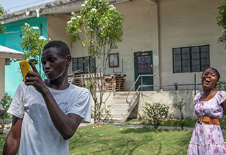 Teenage boy takes a selfie with cellphone camera with teenage girl in the background, in outdoor courtyard in Haiti
