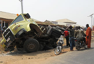 On the side of the road, a badly damaged large truck lays on its side, people gather to inspect the accident.