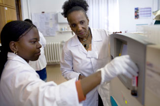 Two female researchers working in a lab, younger one works with equipment, older one by her side observes