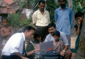 Outdoors, child seated at laptop is instructed by man in shirt and tie, adult and another child look on close by, more men and c