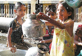 Three young girls gather by water pump, one holds up large metal jug, drops of water splashing