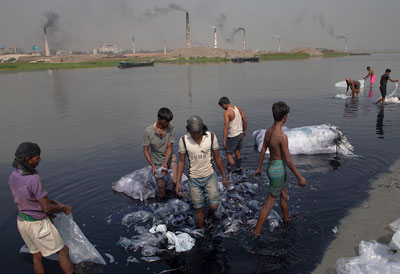 Many men wading in river bank rinsing large clumps of plastic bags, smoking factories in background on opposite bank