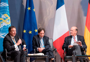 Roger Glass seated on a stage during the World Health Summit with two other men, multiple flags in the background