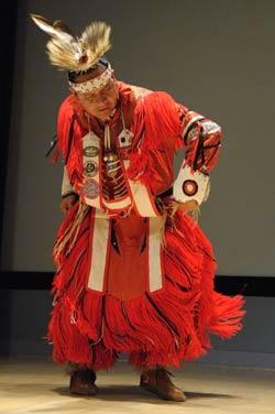 Native American man in traditional dress, including a feathered headdress, a red fringed robe and moccasins, performs dance