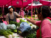 Photo: at outdoor vegetable market woman facing away from camera reaches across a table full of leafy green vegetables