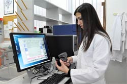 Woman in lab coat in front of computer screen uses hand-held scanner attached to desktop computer.