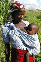 Photo: A woman in a head wrap stands in a field, a toddler in a sling at her side breastfeeds