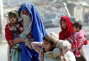 Two Pakistani women with colorful scarves wrapped around their heads walk carrying toddlers, older boy walks beside them