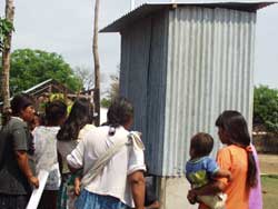 Many people, including women holding toddlers, gather around a small corrugated metal outhouse