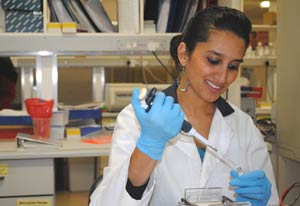Photo: Raabya Rossenkhan in white lab coat and blue latex gloves, smiling, lab in background
